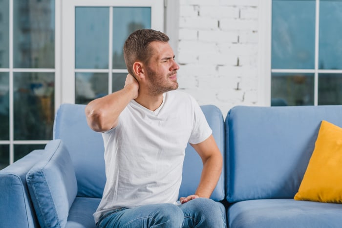 Man sitting on a couch, holding his neck in discomfort.