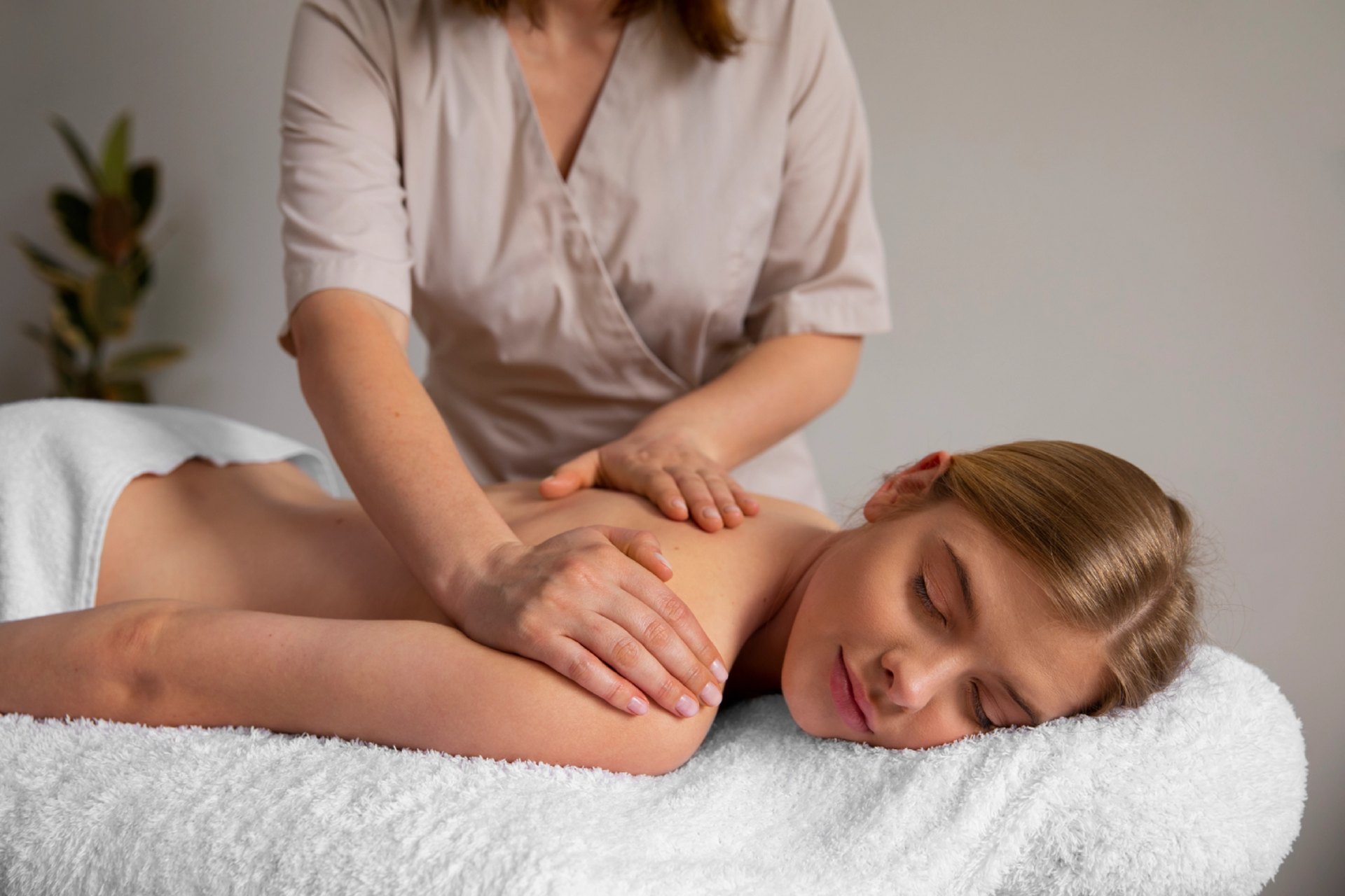 A therapist gives a back massage to a blonde woman lying on a white towel.